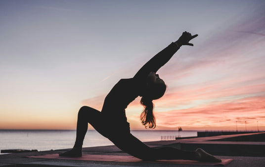 woman in yoga pose at sunrise near water, symbolizing morning wellness routine and recovery